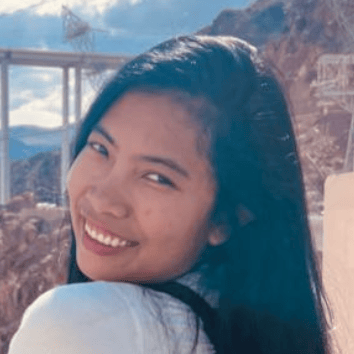 A close-up photo of a woman smiling outdoors with long dark hair, wearing a white shirt. In the background, there are mountains and part of a bridge structure. The image captures a bright, sunny day.